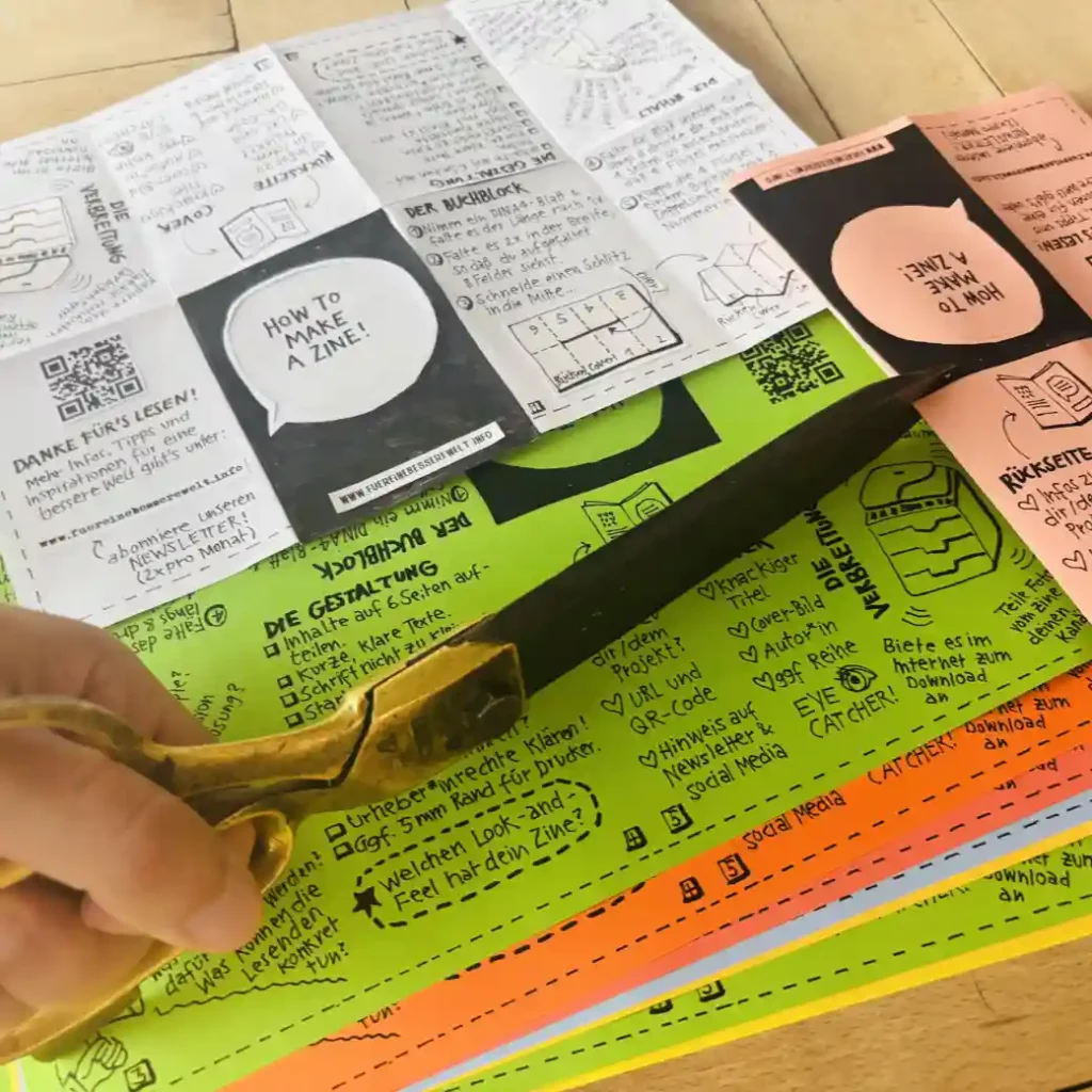Mehrere kopierte Vorlagen, die zu kleinen Zines gefaltet werden sollen. Eine Hand hält eine Schere, um den Schlitz in die Mitte zu schneiden.