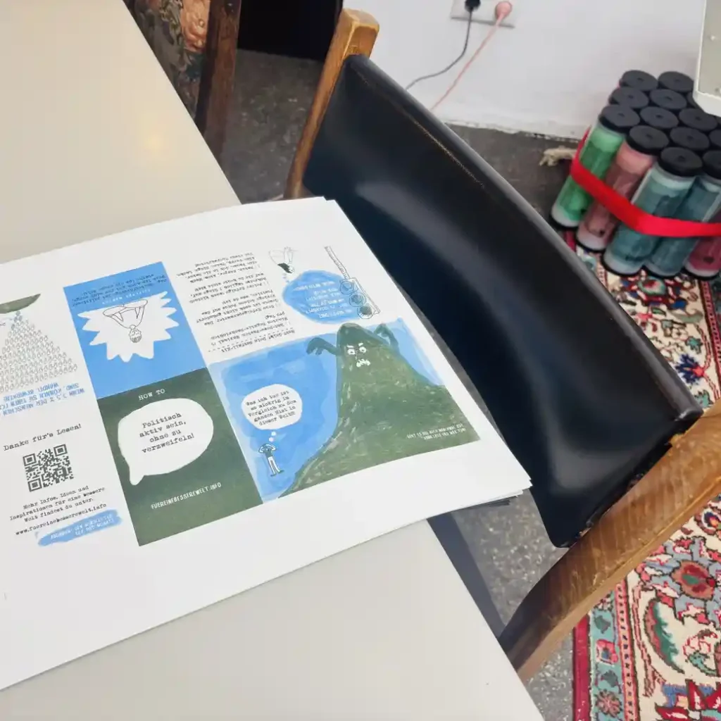 Ein Stapel bedruckter Zine-Bögen auf einem Tisch, daneben ein Stuhl und im Hintergrund mehrere Risoprint-Farbpatronen.